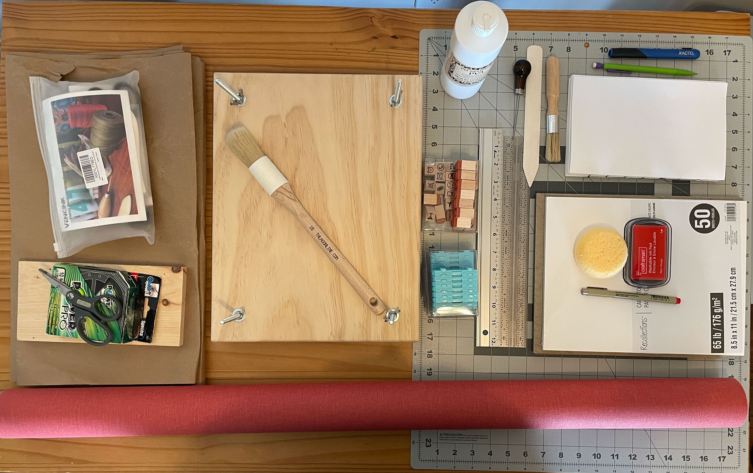 Various craft and bookbinding tools, including a book press, cutting mat, boards and buckram cloth, glue, brushes, stamps, rulers, thread, bonefolder, and awl.